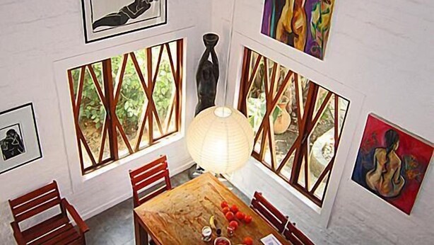 A dining area with a wooden table, chairs, and a view through the windows. There are artworks on the walls and a lamp on the table. - Alternative Space B & B (Swakopmund)