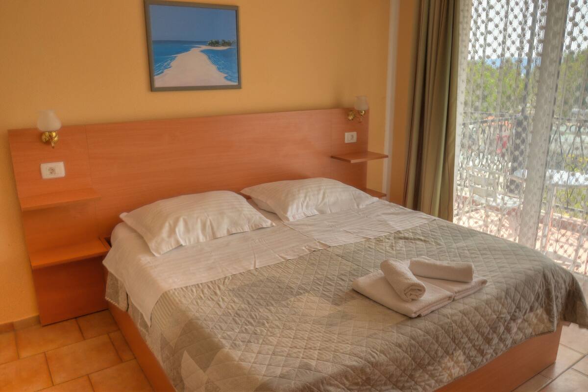 A double bed with a quilted bedspread and two pillows, a nightstand with a lamp, and a painting of a beach scene on the wall.