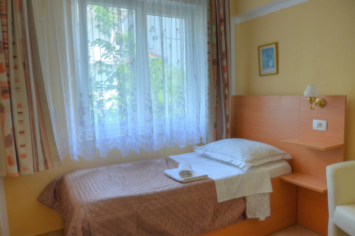 A neatly made bed with a white pillow and a folded towel, a bedside table with a lamp, and a window with sheer curtains.
