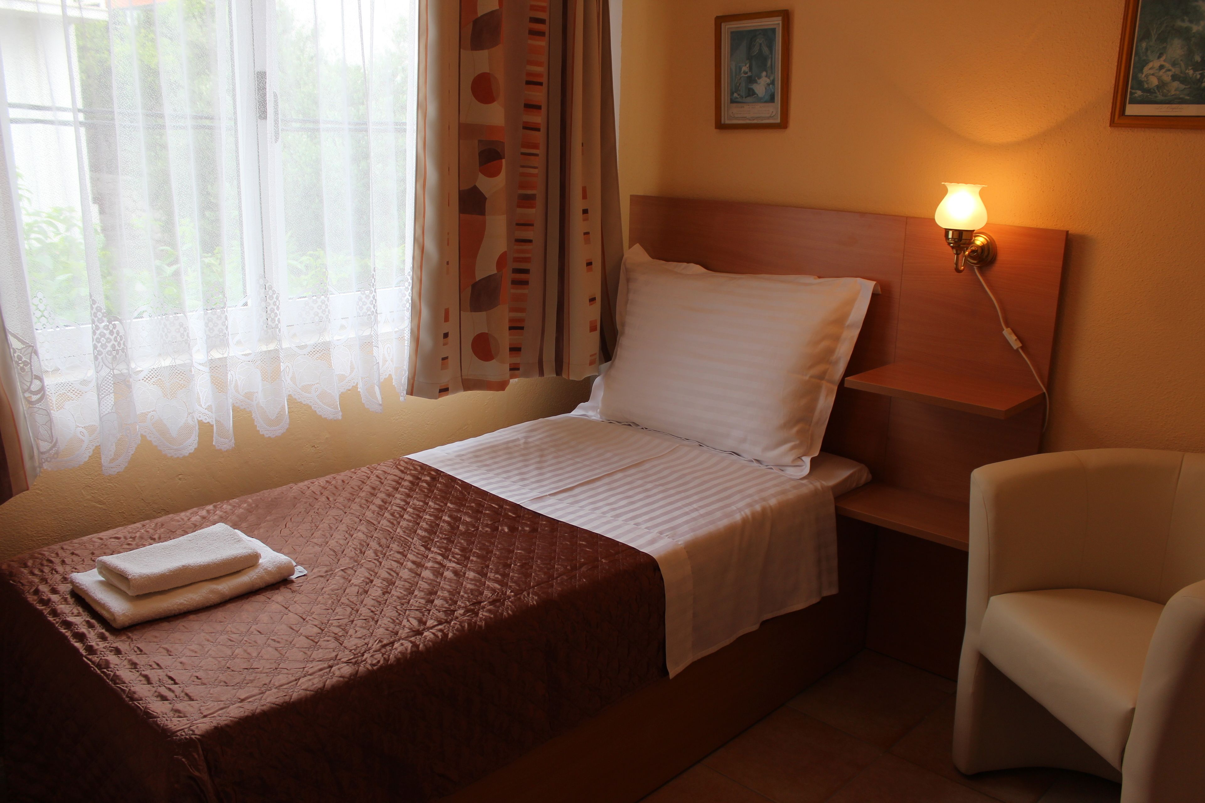 A single bed with a brown quilt, a white pillow, and two folded towels. There is a bedside lamp on the nightstand.