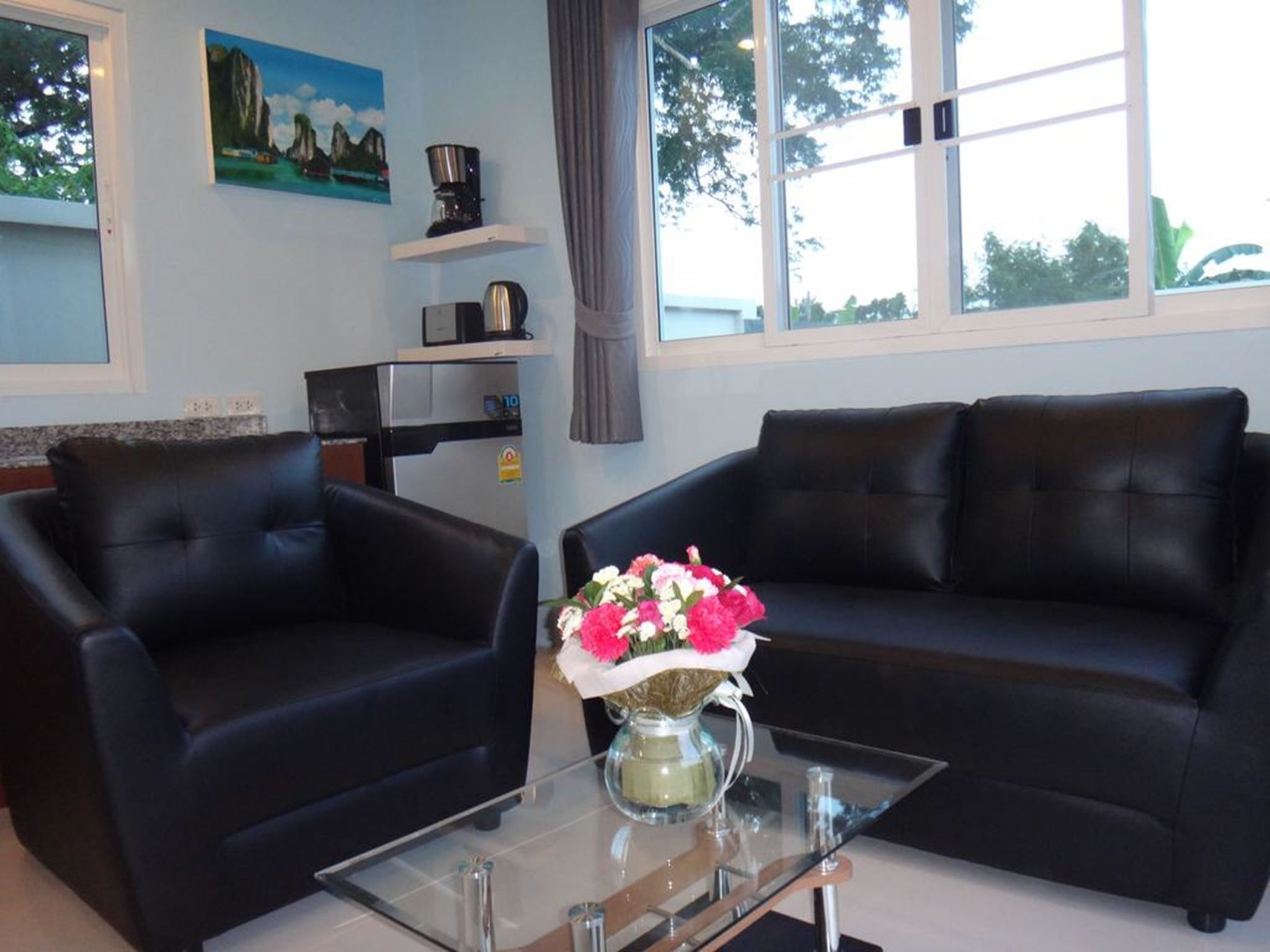 Ein Wohnzimmer mit Glastisch, zwei schwarzen Ledersofas, einem Gemälde an der Wand und einer Küchenzeile inklusive Kühlschrank und Kaffeemaschine.