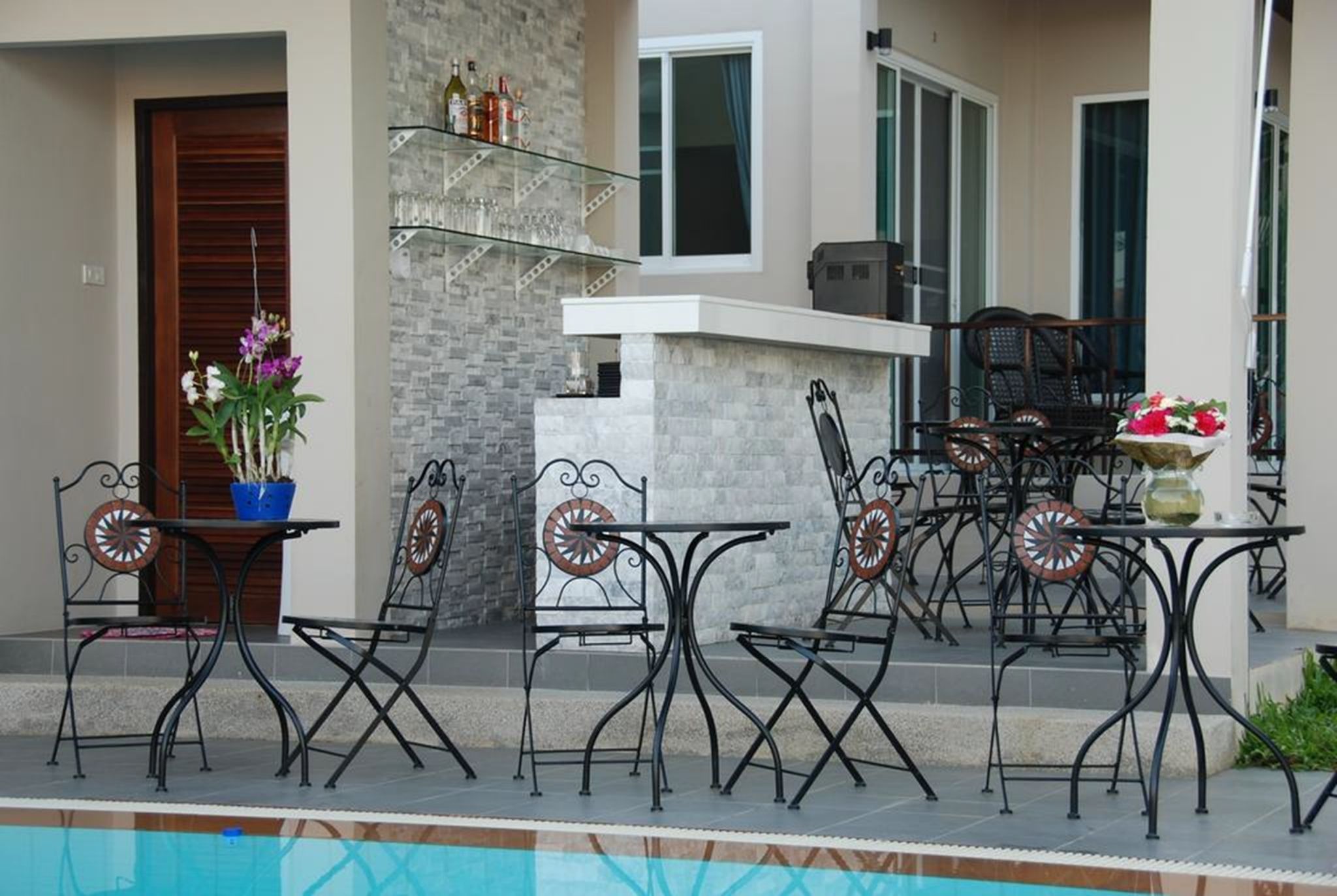 An outdoor patio with a pool, featuring a bar area with a stone wall, metal chairs, and a table with a potted plant.