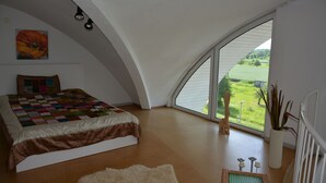 Een slaapkamer met een groot bed, uitzicht op een groen landschap door een raam en een wit plafond.