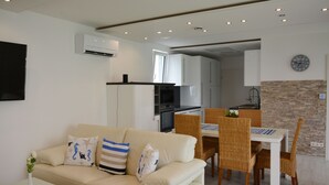 Een moderne woonkamer met een beige bank, een eettafel met rieten stoelen, een keuken met witte kasten en een accentmuur van baksteen.