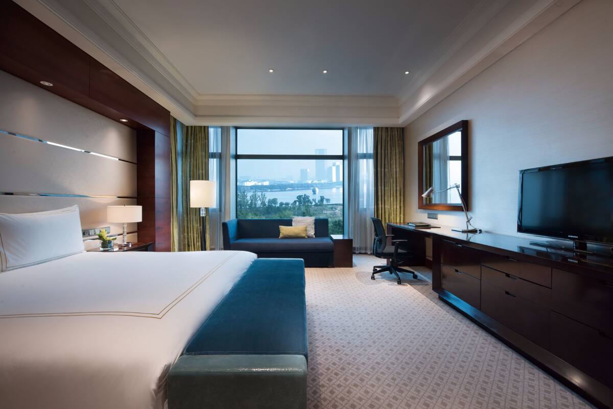 A hotel room with a large bed, a sofa, a desk with a TV, and a view of the city.