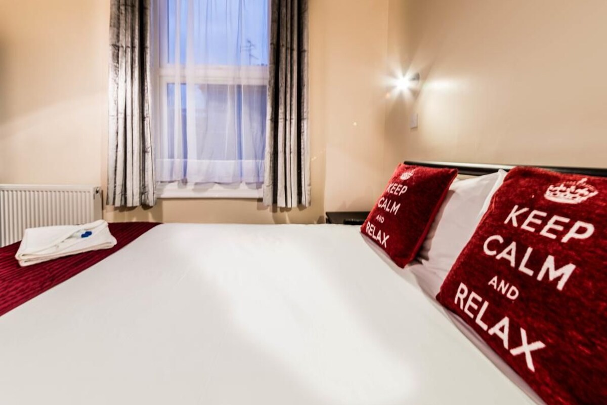 A hotel room with a bed, a window with curtains, a radiator, and a red pillow with 'KEEP CALM AND RELAX' text.