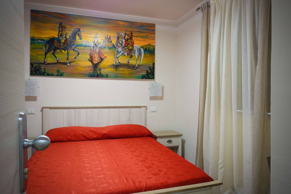 A bedroom with a bed, a nightstand, a painting, and a curtain.