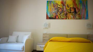 Ein Schlafzimmer mit einem weißen Sofa, einem Bett mit gelber Tagesdecke und einem farbenfrohen Bild an der Wand.