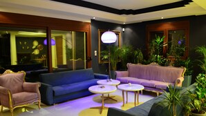 A modern living room with a blue sofa, a pink sofa, and a gold armchair. There are two round tables with white tops and gold legs. The room has large windows with a view of the city. - Gold Luxury Villa Ialysos (Rhodes)