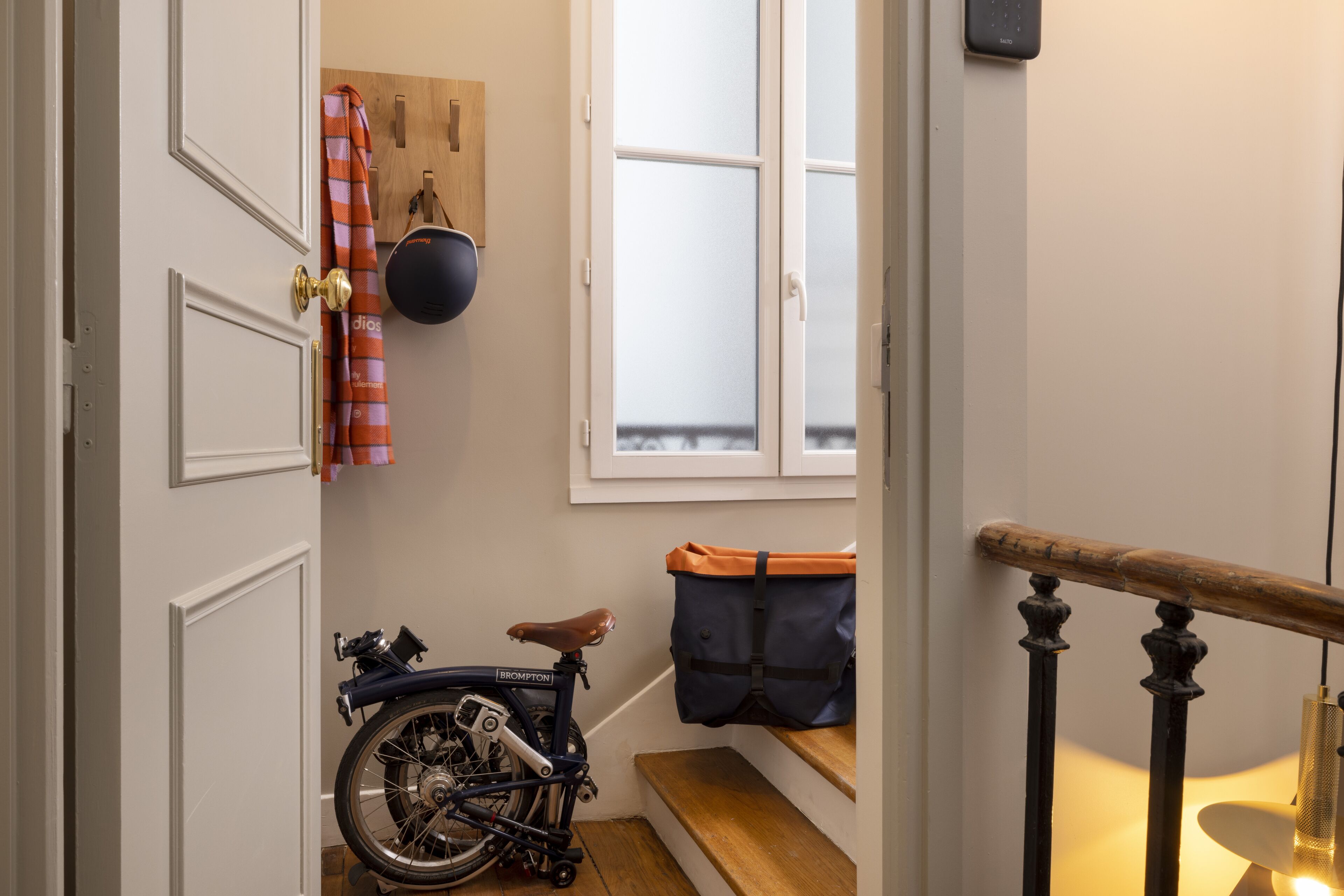 A narrow hallway with a bicycle parked by a door, a window with frosted glass, and a wooden bench with a bag on it.
