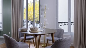 A dining area with a round table, four grey chairs, a gold floor lamp, and a view of a balcony with yellow foliage. - Yuna Blanche - Serviced Apartments (Paris)