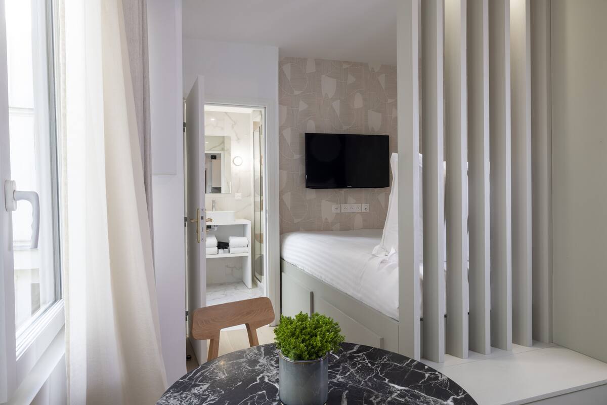 A modern hotel room with a bed, a TV mounted on the wall, a bathroom visible through an open door, and a small table with a plant.