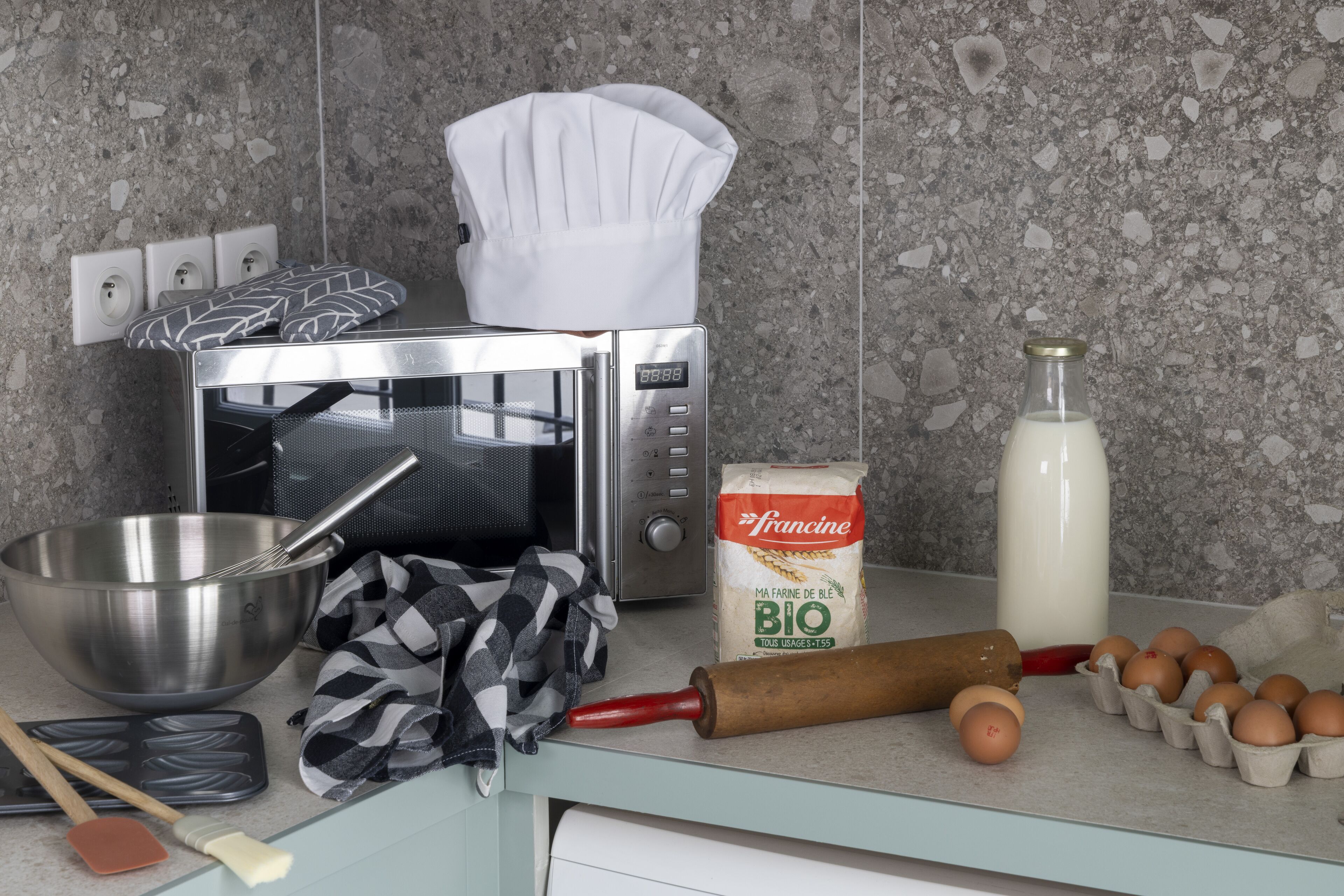 A kitchen counter with a microwave, a chef's hat, a whisk, a rolling pin, eggs, a bottle of milk, a bag of flour, and a spatula.