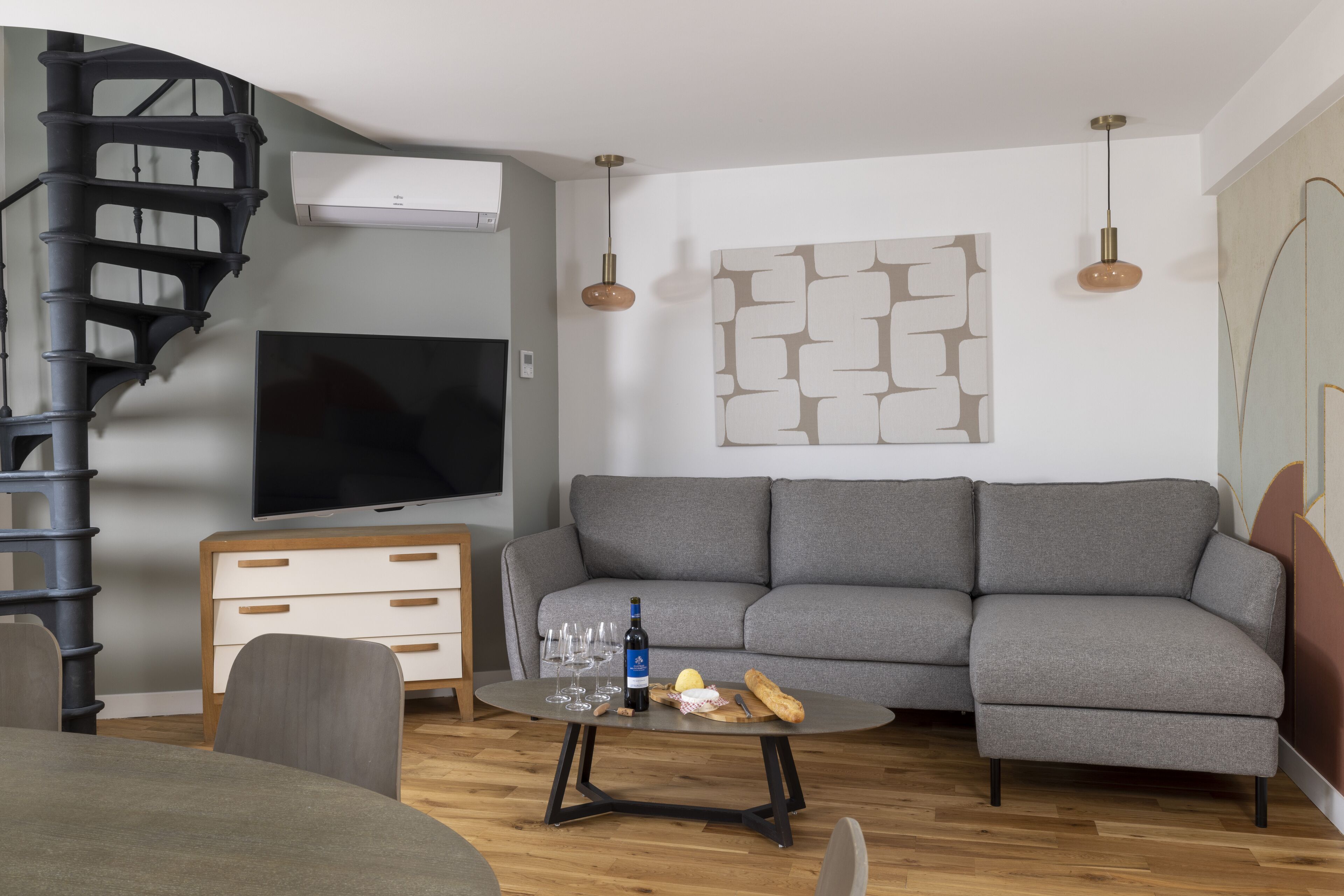 A modern living room with a grey sofa, a TV, a small round table with a bottle and glasses, a spiral staircase, and a wall-mounted air conditioner.