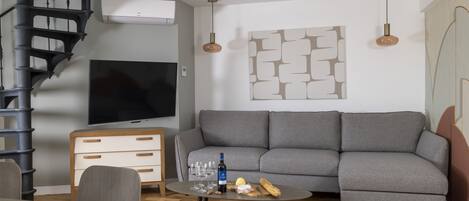 A modern living room with a grey sofa, a TV, a small round table with a bottle and glasses, a spiral staircase, and a wall-mounted air conditioner.