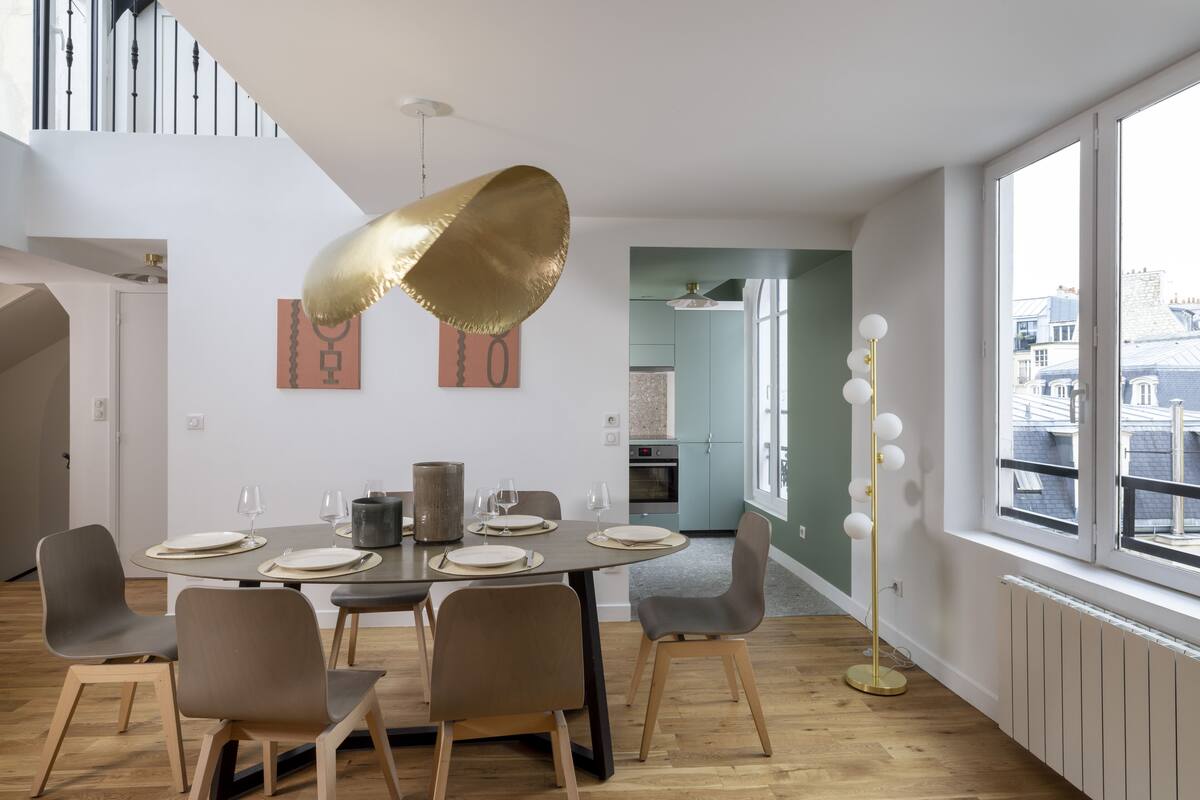 A modern dining area with a round table set for four, a gold pendant light, and a view of the city through large windows.