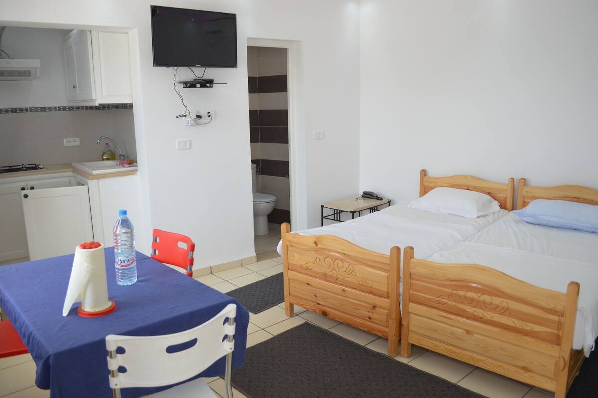 A bedroom with a wooden bed, a small table with a chair, a TV mounted on the wall, and a kitchenette with a sink and stove.