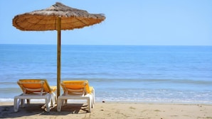 Une plage avec deux chaises longues blanches sous un parasol en chaume, donnant sur l’océan.