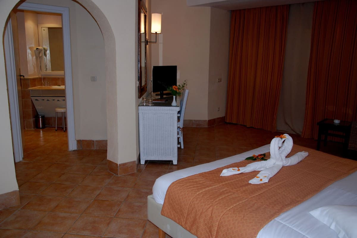 A hotel room with a bed, a desk, a chair, a mirror, a lamp, and a bathroom visible through an archway.