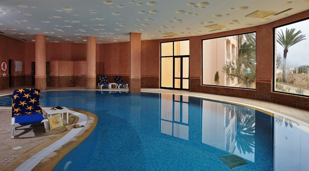 An indoor swimming pool with clear blue water, large windows offering views of palm trees and buildings, and a lounge area with a chair and sun loungers.
