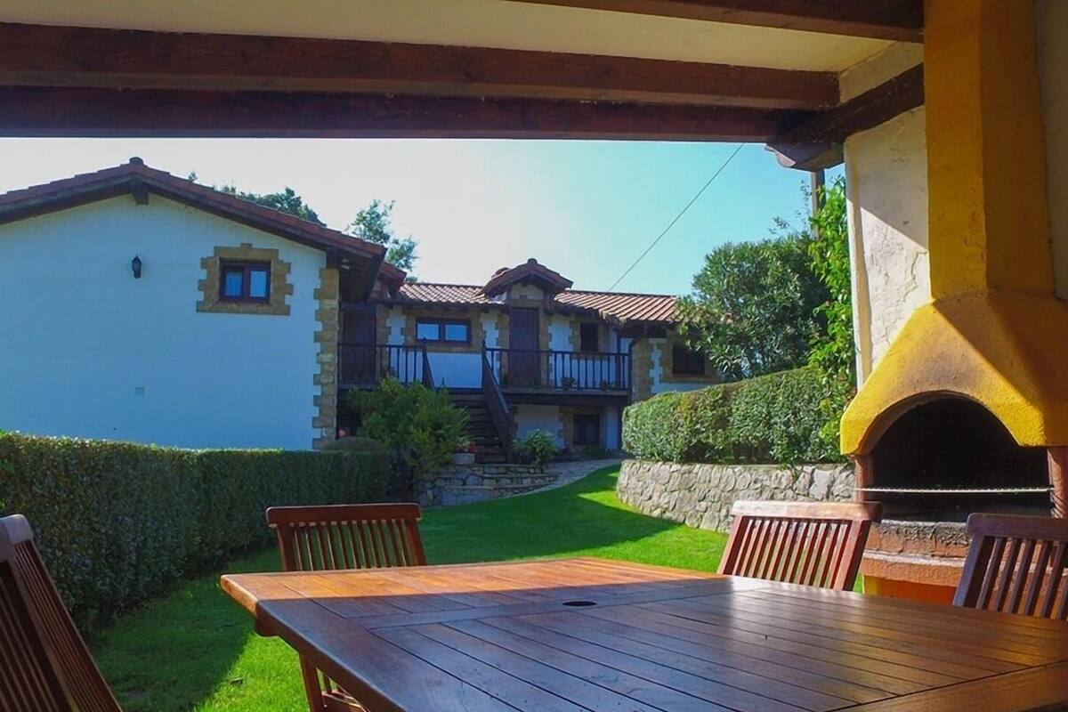 Un patio con mesa y sillas de madera, un muro de piedra y un edificio con balcón al fondo.