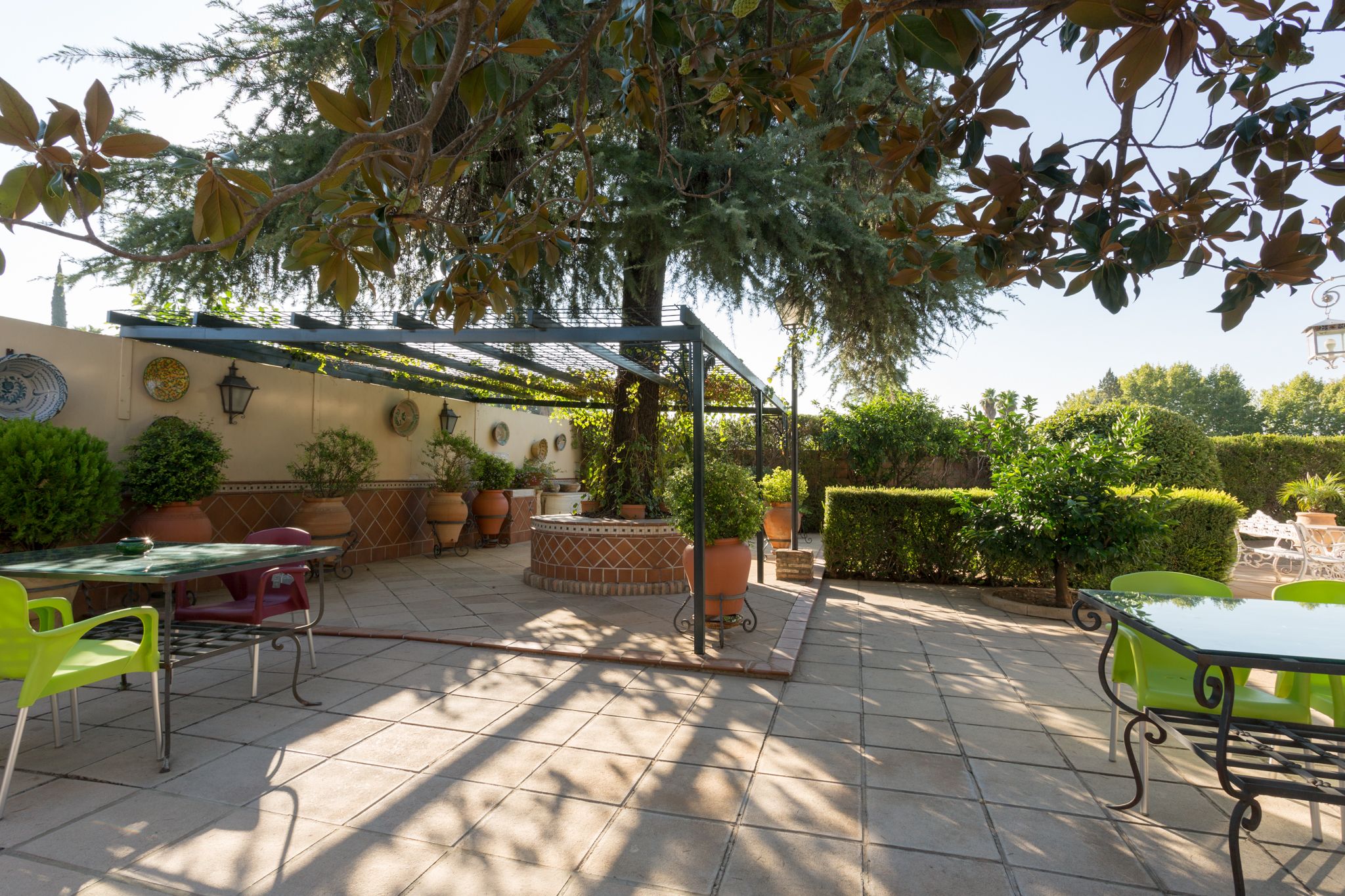 Un patio al aire libre con una pérgola, plantas en macetas y una mesa con sillas.