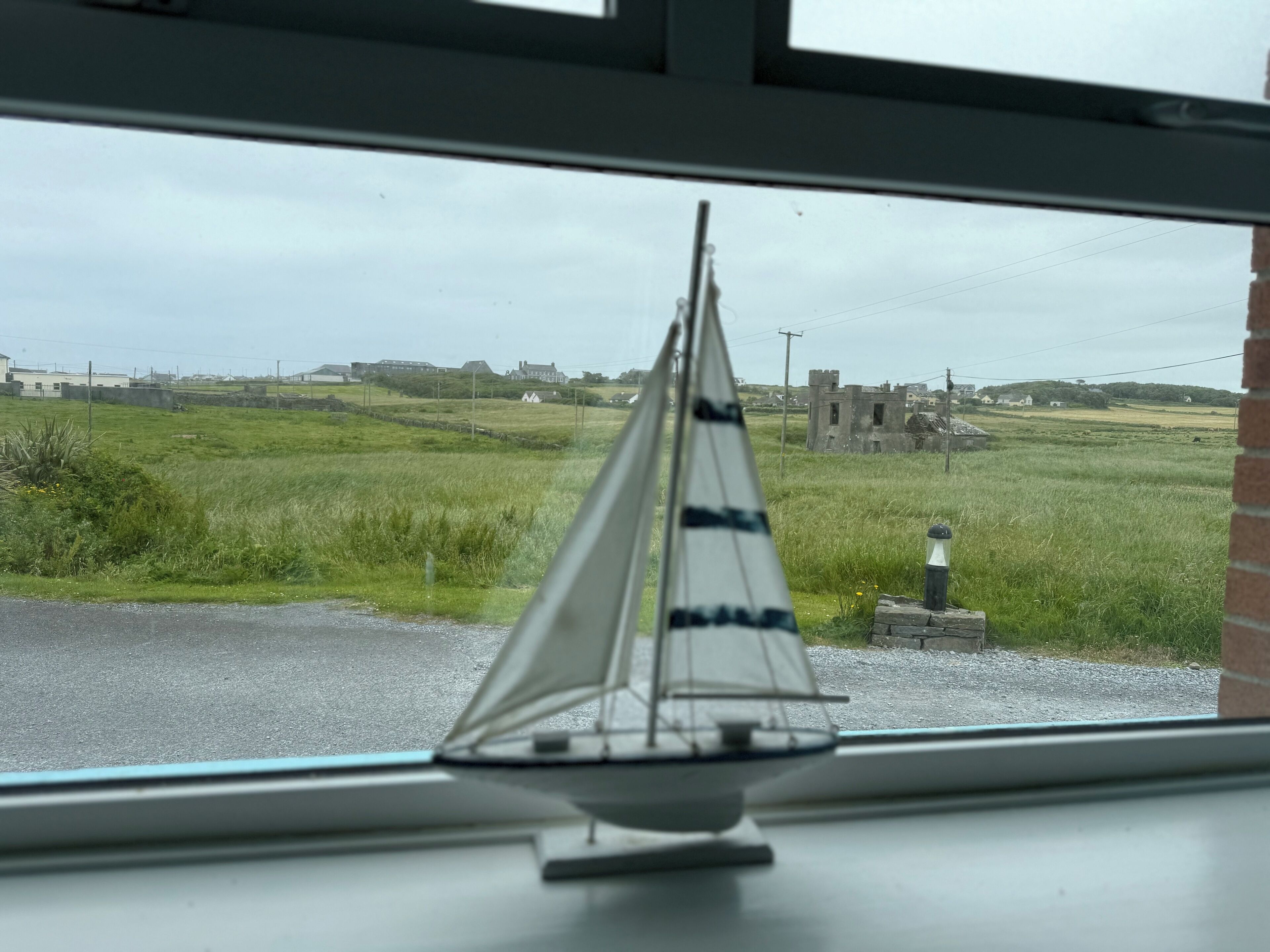 A model sailboat on a windowsill with a view of a grassy field and buildings in the distance.