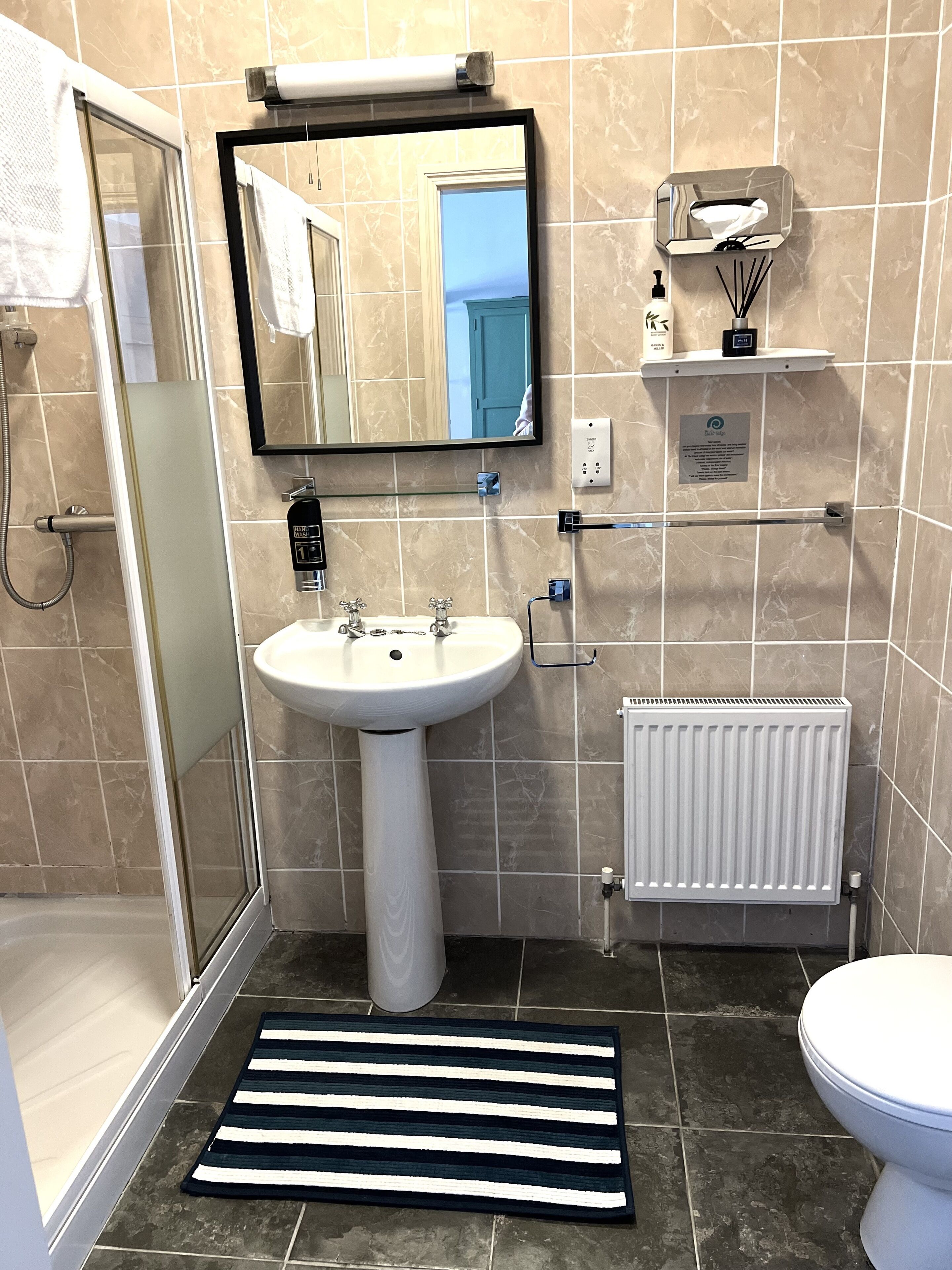 A bathroom with a white pedestal sink, a mirror above it, a towel rack, a shower area, a toilet, and a radiator.