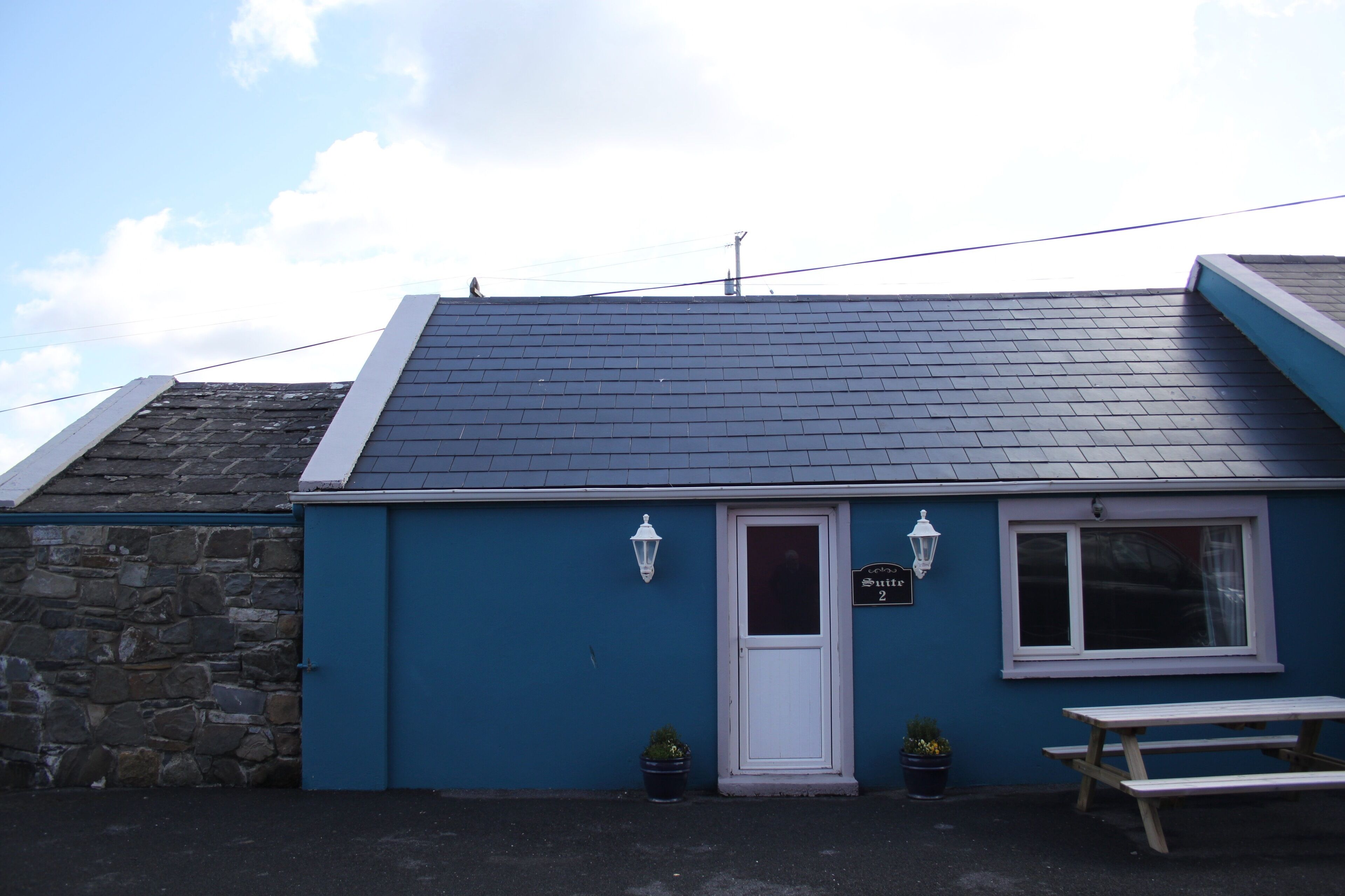 A small building with a dark blue exterior, a white door, and a window. There is a sign above the door, a bench outside, and two potted plants.