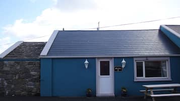 A small building with a dark blue exterior, a white door, and a window. There is a sign above the door, a bench outside, and two potted plants.