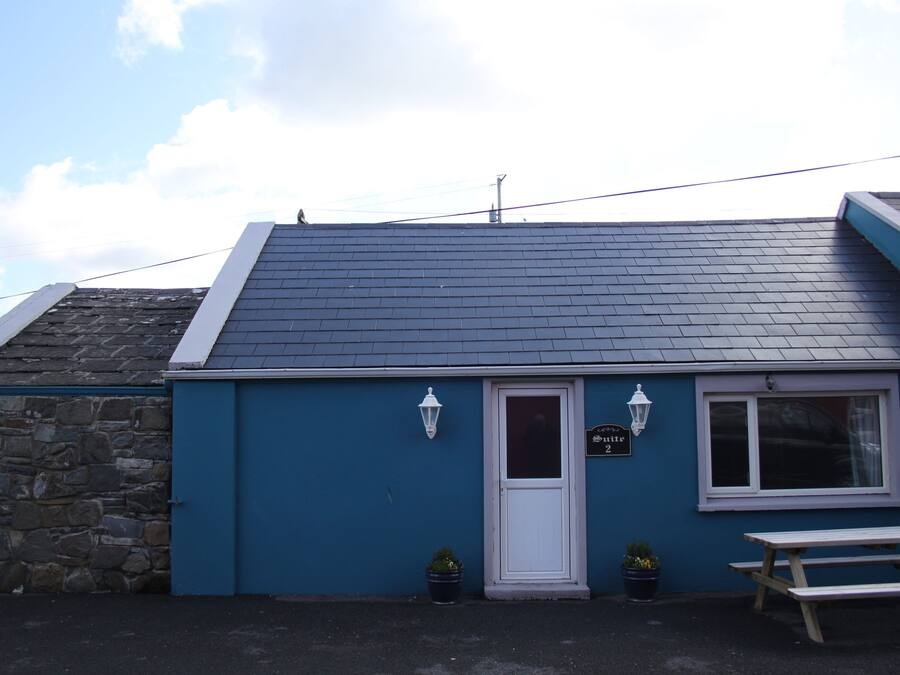 A small building with a dark blue exterior, a white door, and a window. There is a sign above the door, a bench outside, and two potted plants.