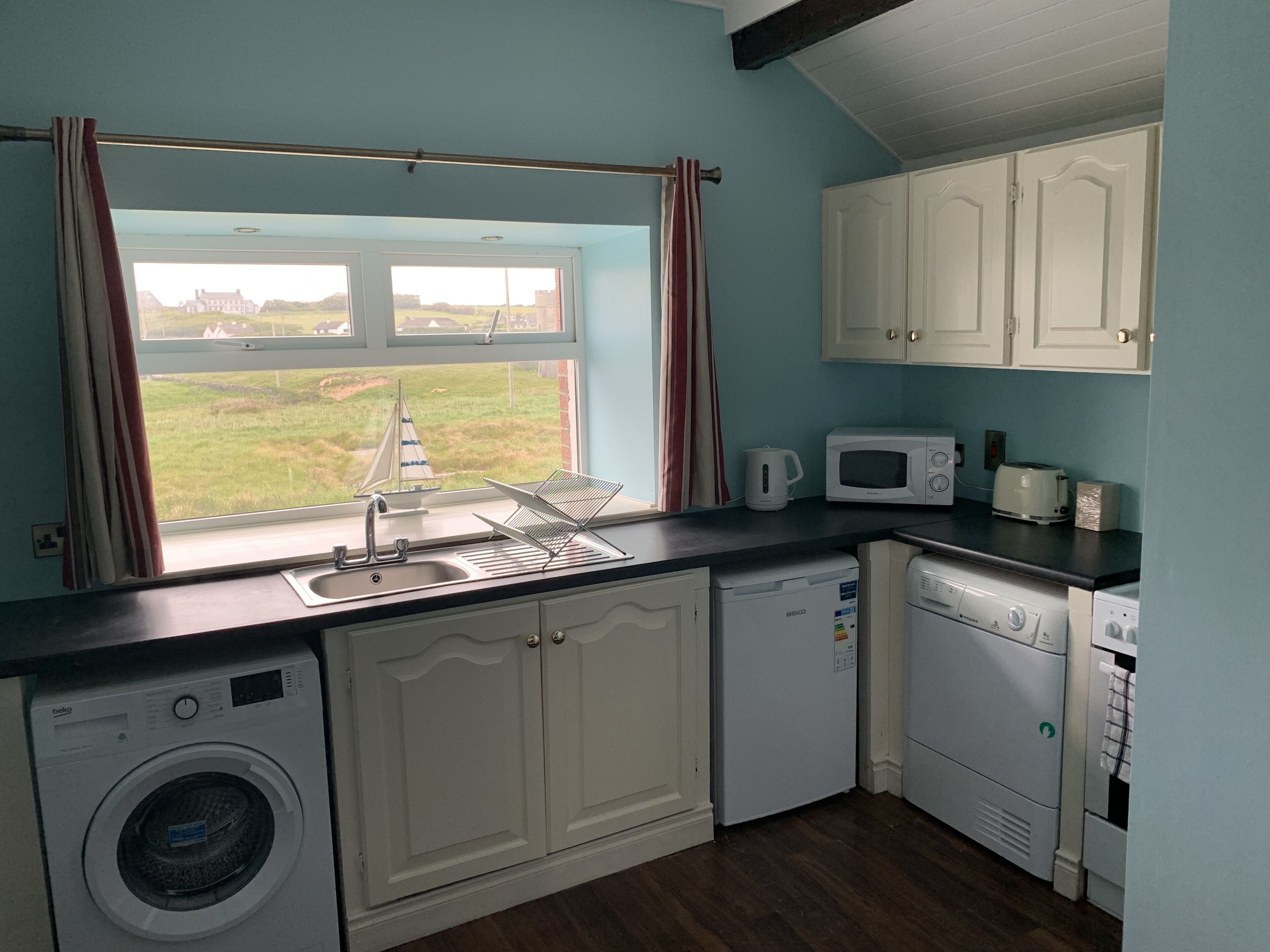 Cocina funcional con lavadora, microondas y lavavajillas. Una ventana con cortinas ofrece vistas al exterior.