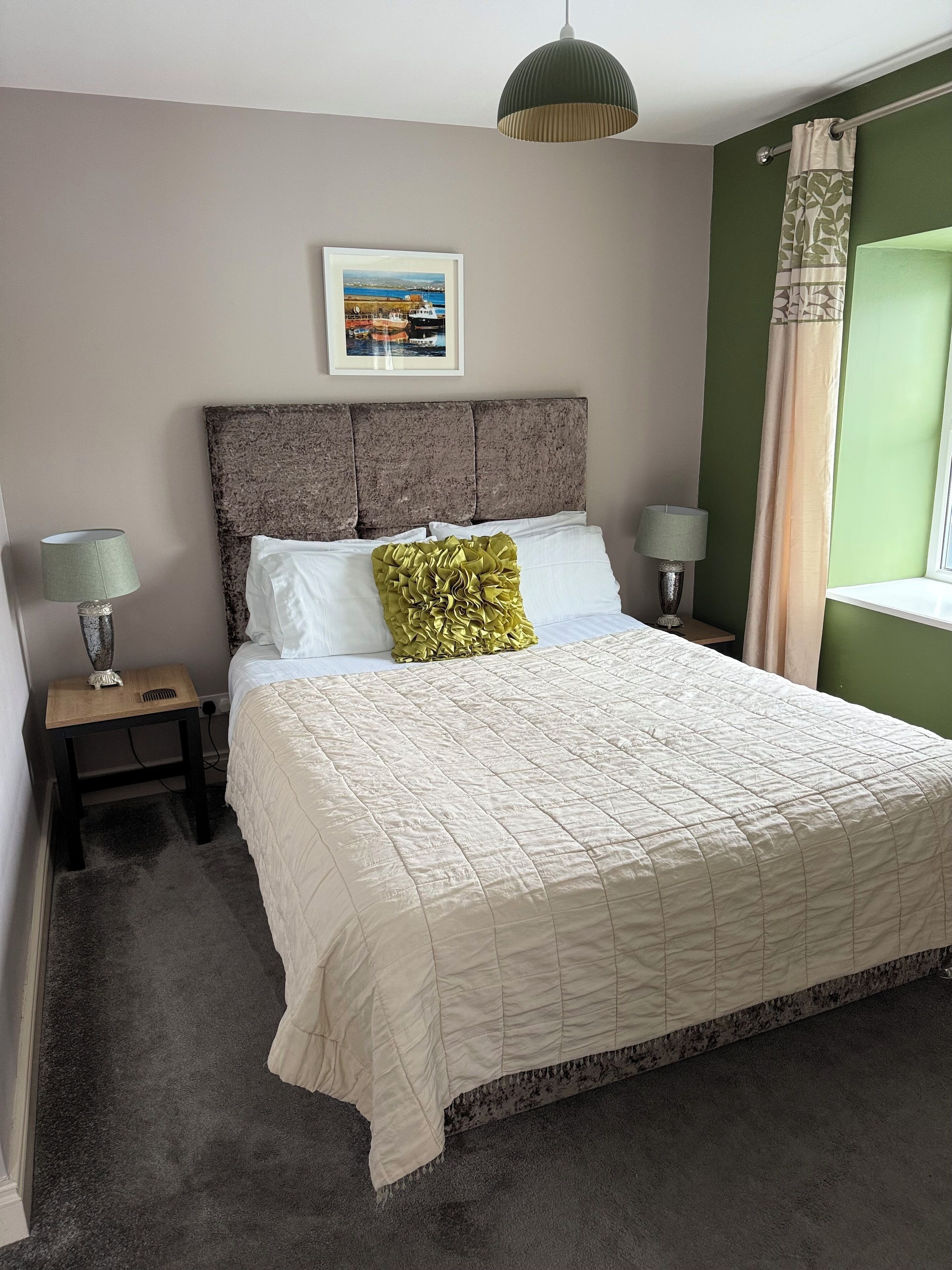 Een netjes opgemaakt bed met een witte quilt, een groen sierkussen en een ingelijst schilderij aan de muur.