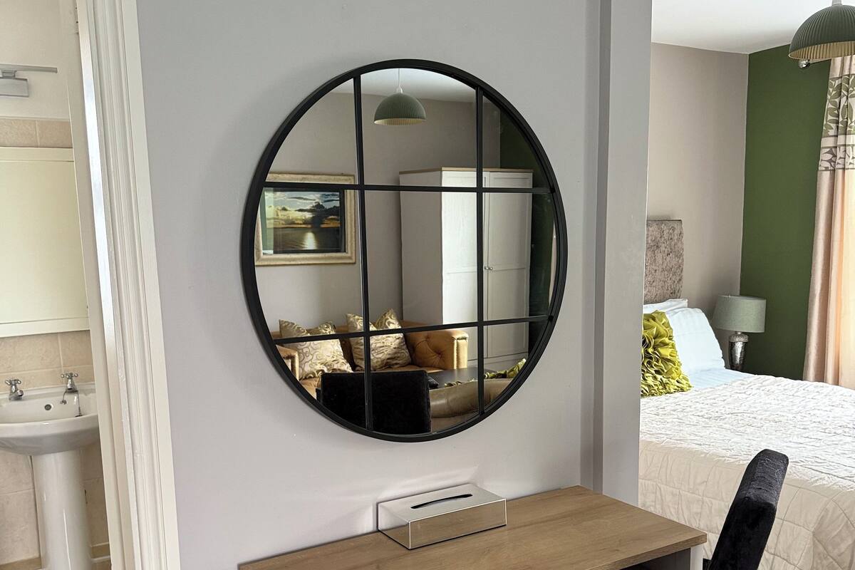 Une chambre avec un miroir rond, un bureau en bois, une chaise noire, un lit avec une couette blanche et une table de chevet avec une lampe.
