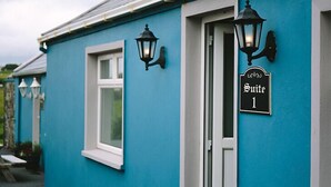 Un bâtiment à la façade turquoise, avec une porte blanche et deux lampes noires fixées au mur. On peut voir une enseigne indiquant « Suite 1 ».
