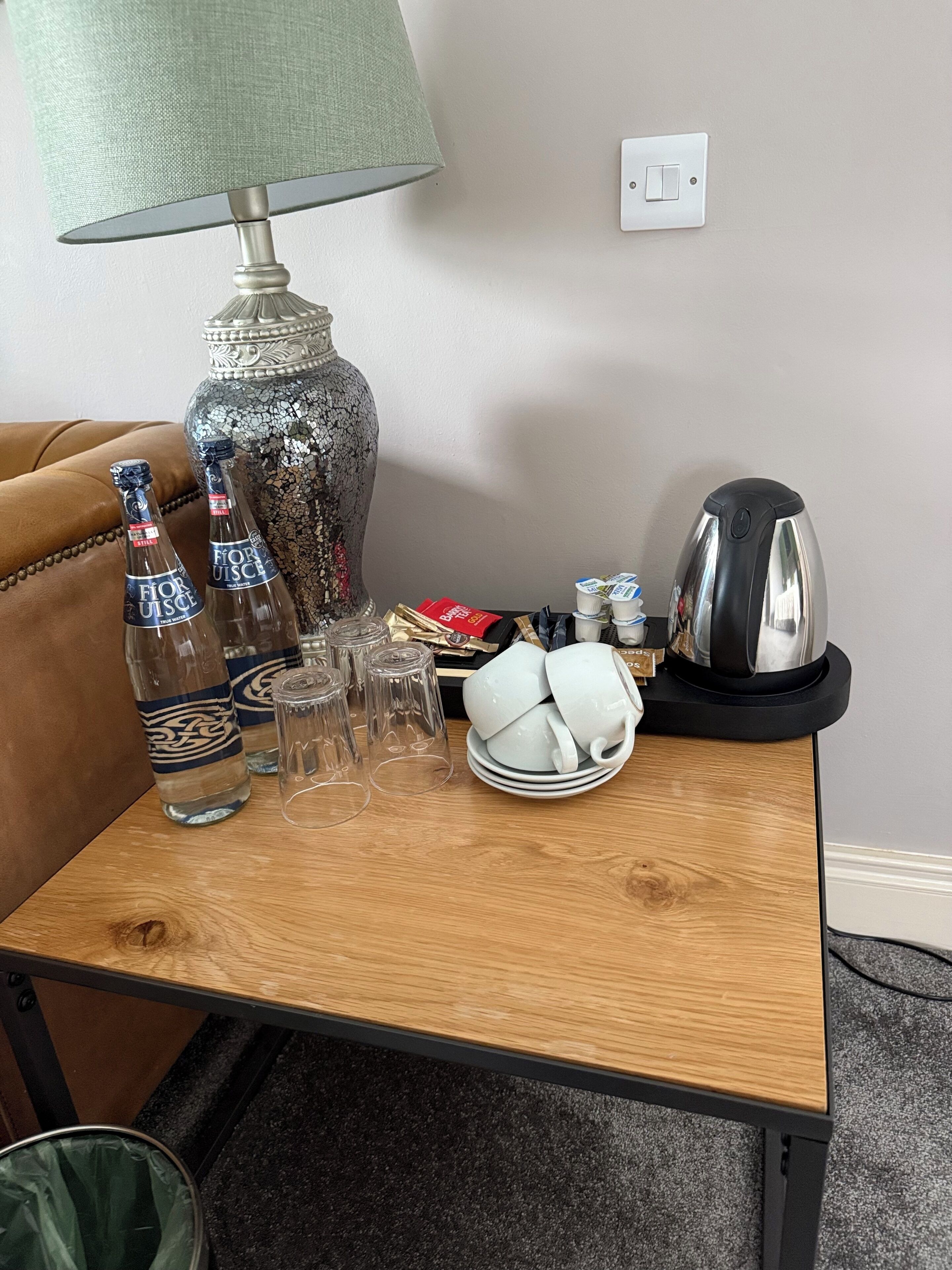 A wooden table with a lamp, two bottles, glasses, a decorative vase, a kettle, and a plate with a cup.