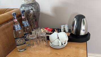 A wooden table with a lamp, two bottles, glasses, a decorative vase, a kettle, and a plate with a cup.