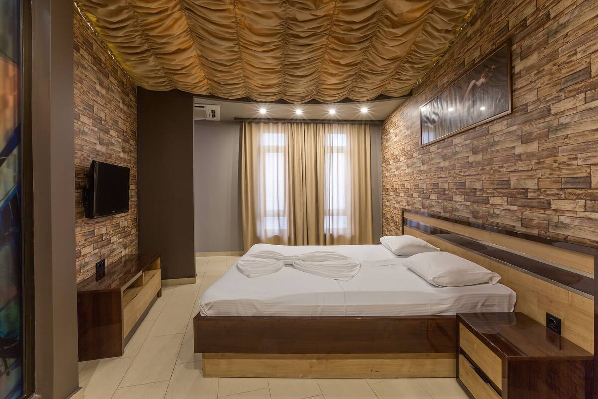 Un dormitorio moderno con cabecera de madera, televisor de pantalla plana montado en una pared de ladrillo, un banco de madera y un ventanal con cortinas.