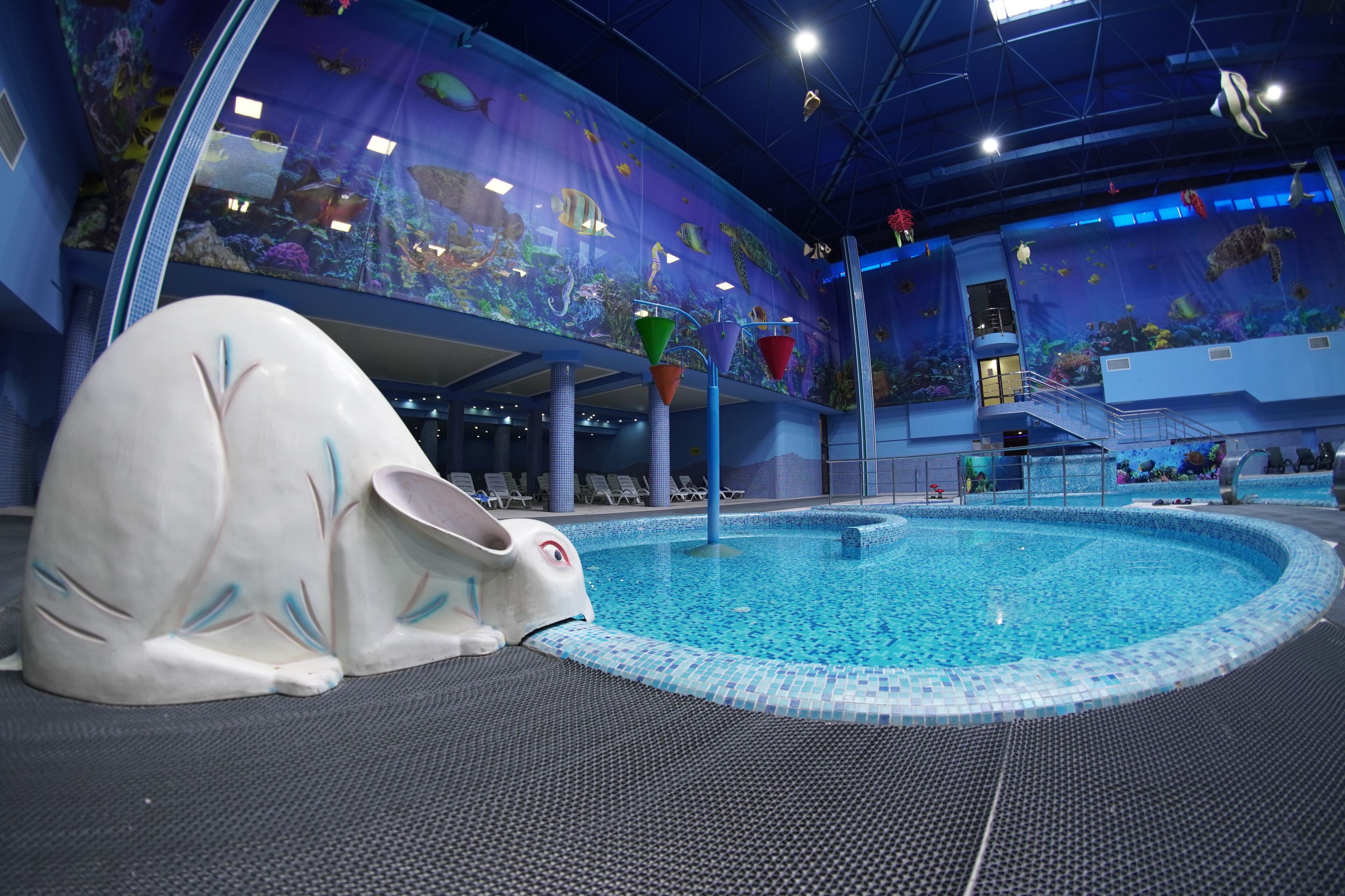 An indoor swimming pool with a large aquarium mural, a dolphin statue, and a seating area with a view of the pool.