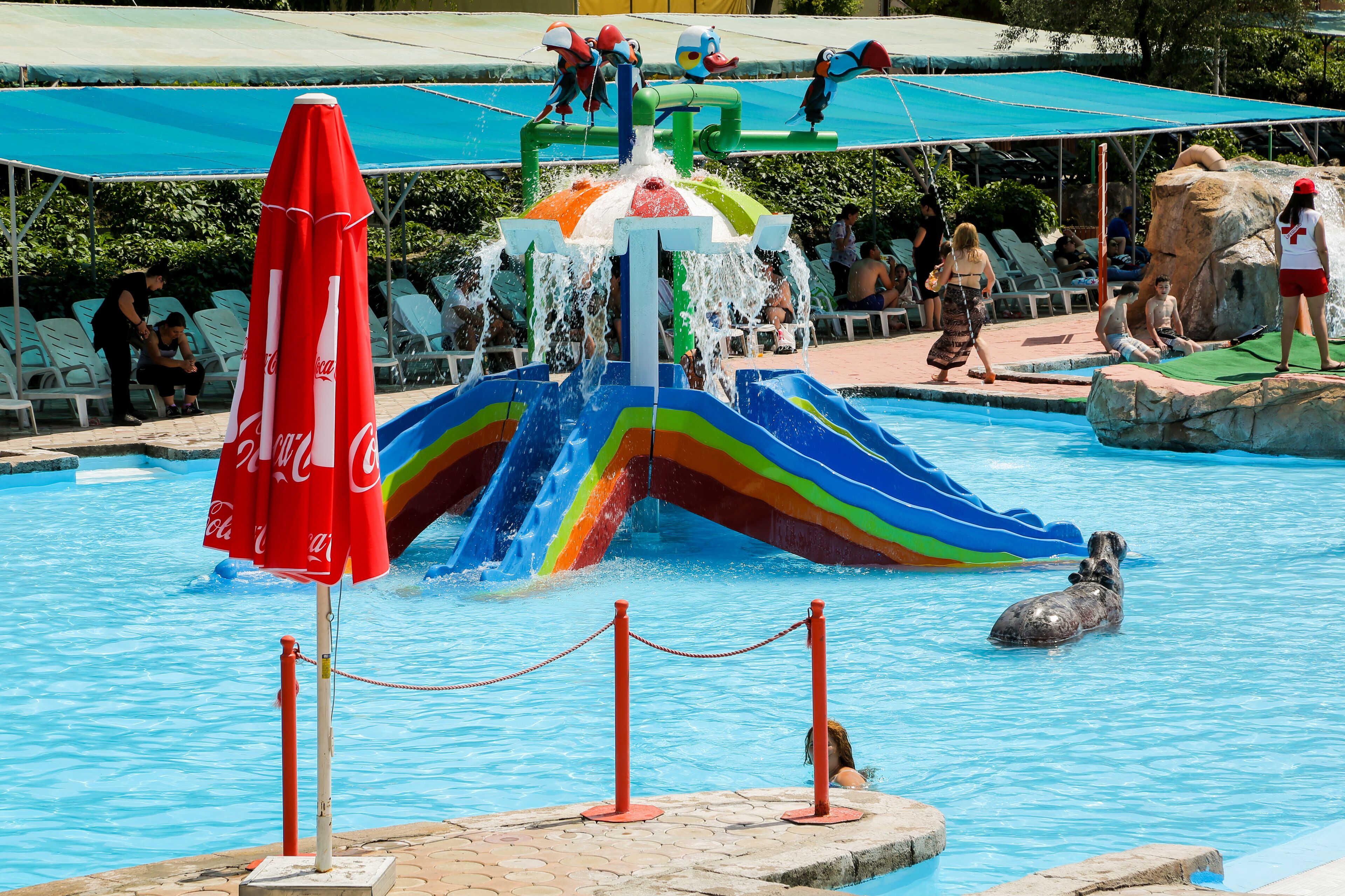 A swimming pool with a water slide and a Coca-Cola branded umbrella.