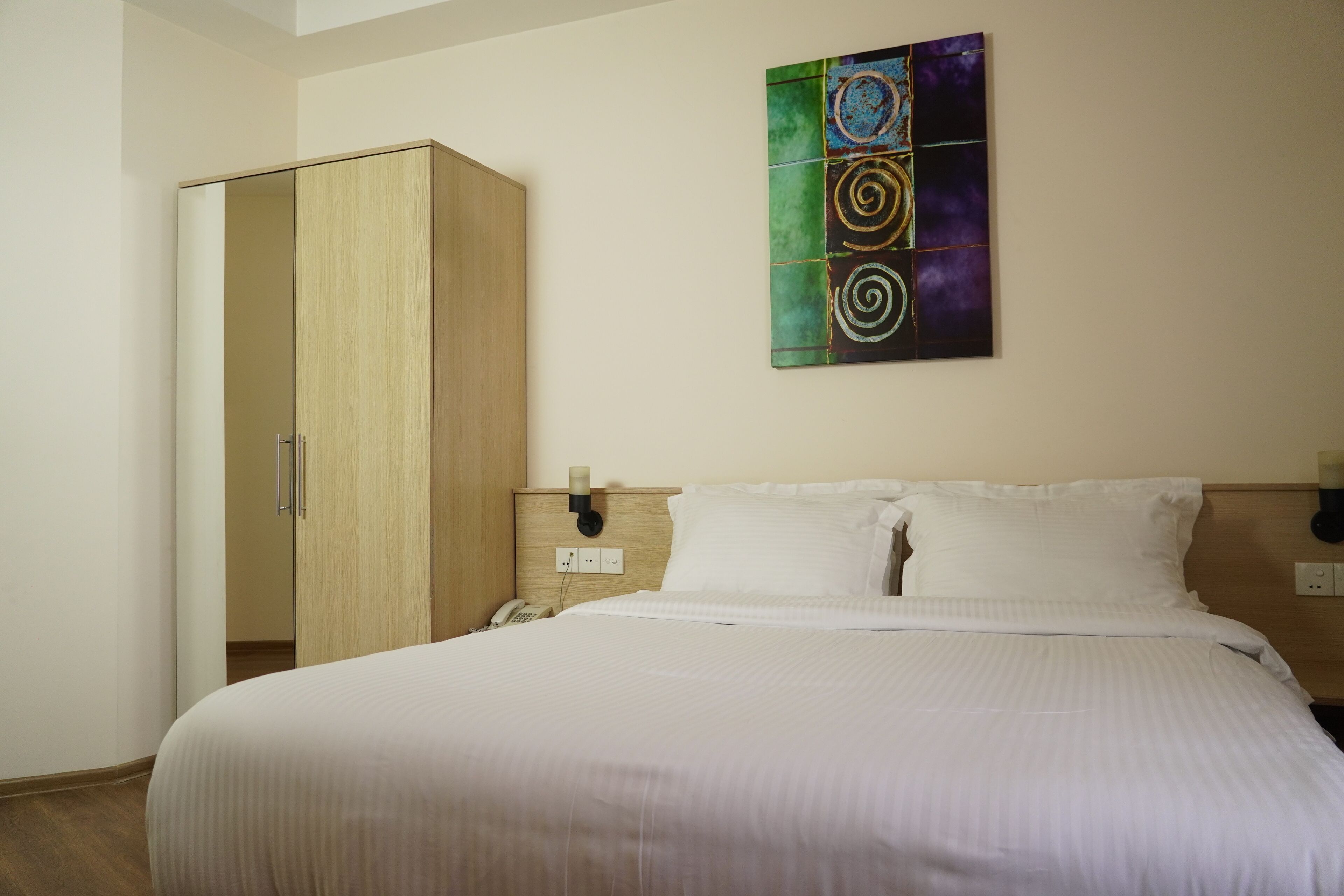 A hotel room with a bed, a wardrobe, a painting on the wall, and a nightstand with a phone.