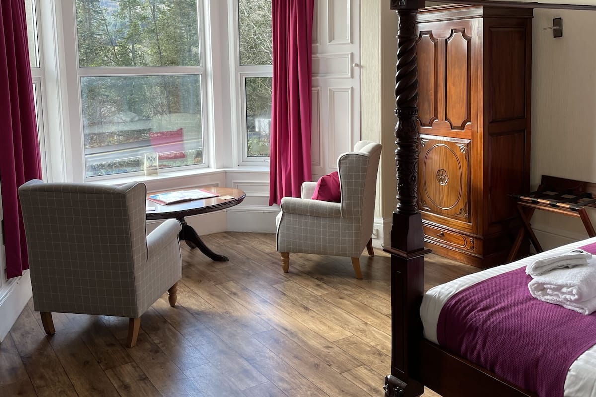 Un dormitorio con cama con dosel, un armario de madera, un escritorio con silla y vistas al exterior a través de amplios ventanales.