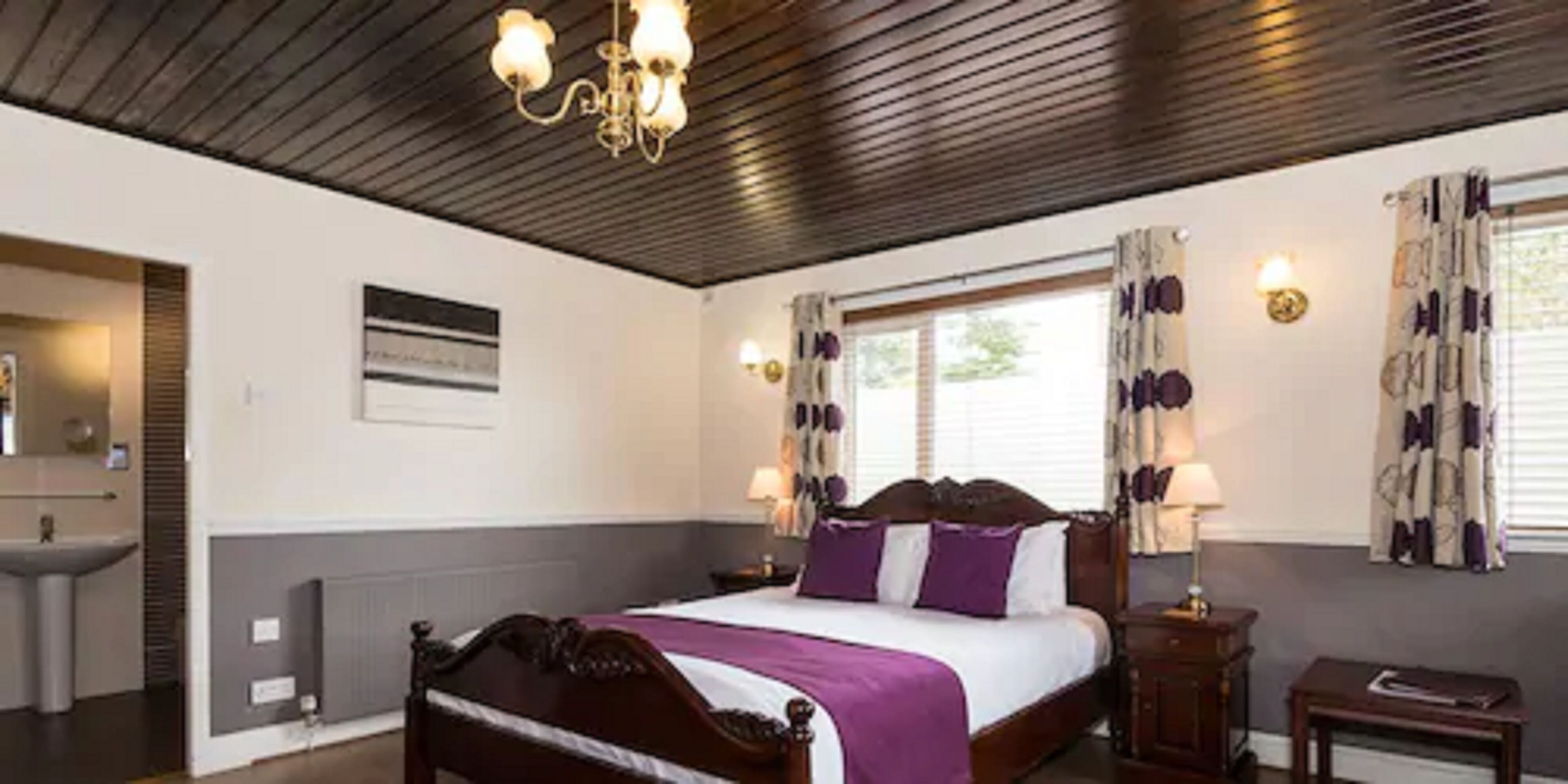 A hotel room with a wooden bed, a bedside table, a nightstand, a window with curtains, a wall-mounted light, and a radiator.