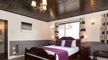 A hotel room with a wooden bed, a bedside table, a nightstand, a window with curtains, a wall-mounted light, and a radiator.