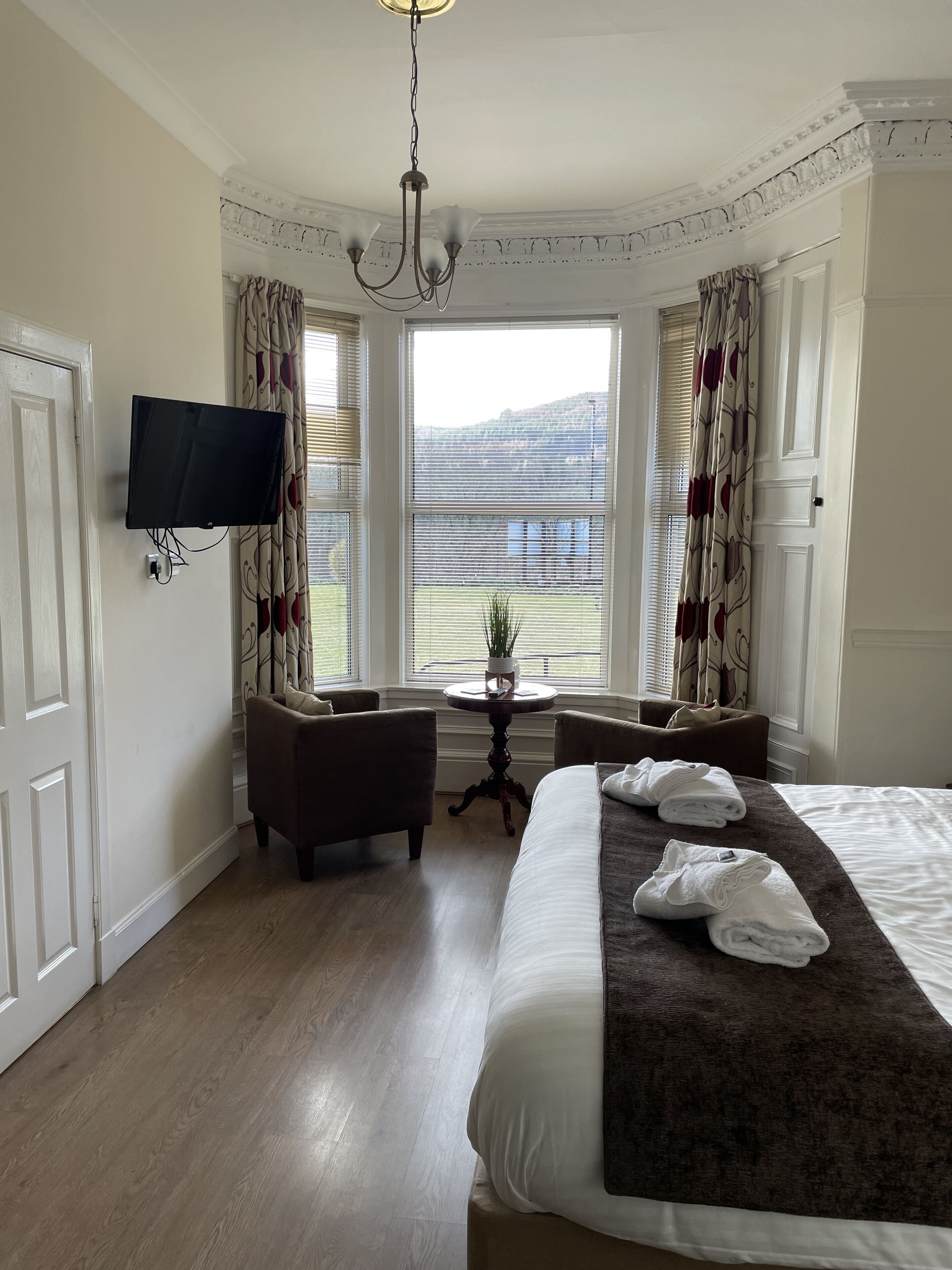 A hotel room with a large bed, a flat-screen TV mounted on the wall, a small table with a plant, two armchairs, and a view of the outdoors through the windows.