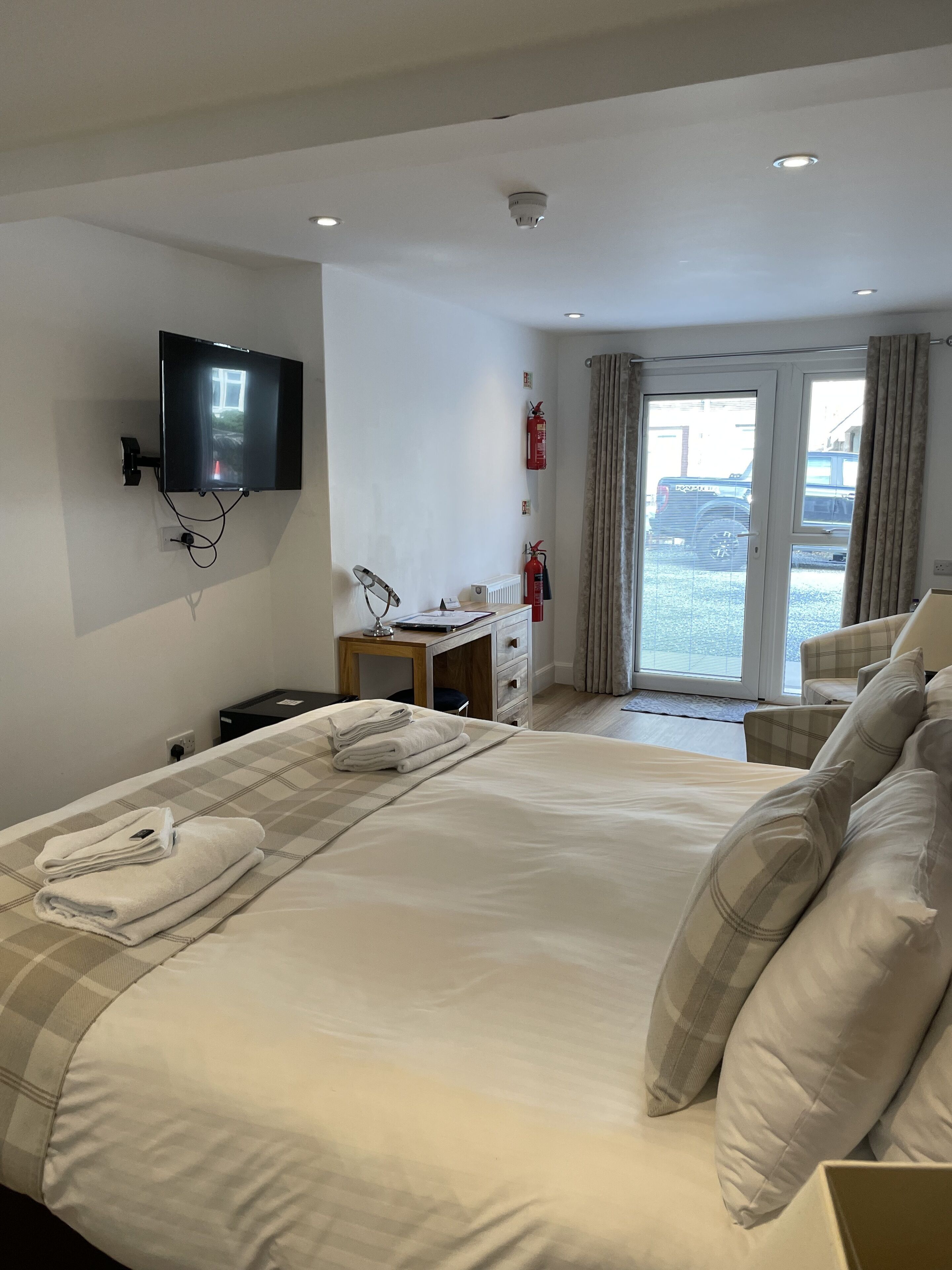 Habitación de hotel con una cama grande, un escritorio, un televisor de pantalla plana montado en la pared, un extintor y vistas al exterior a través de una puerta corredera.