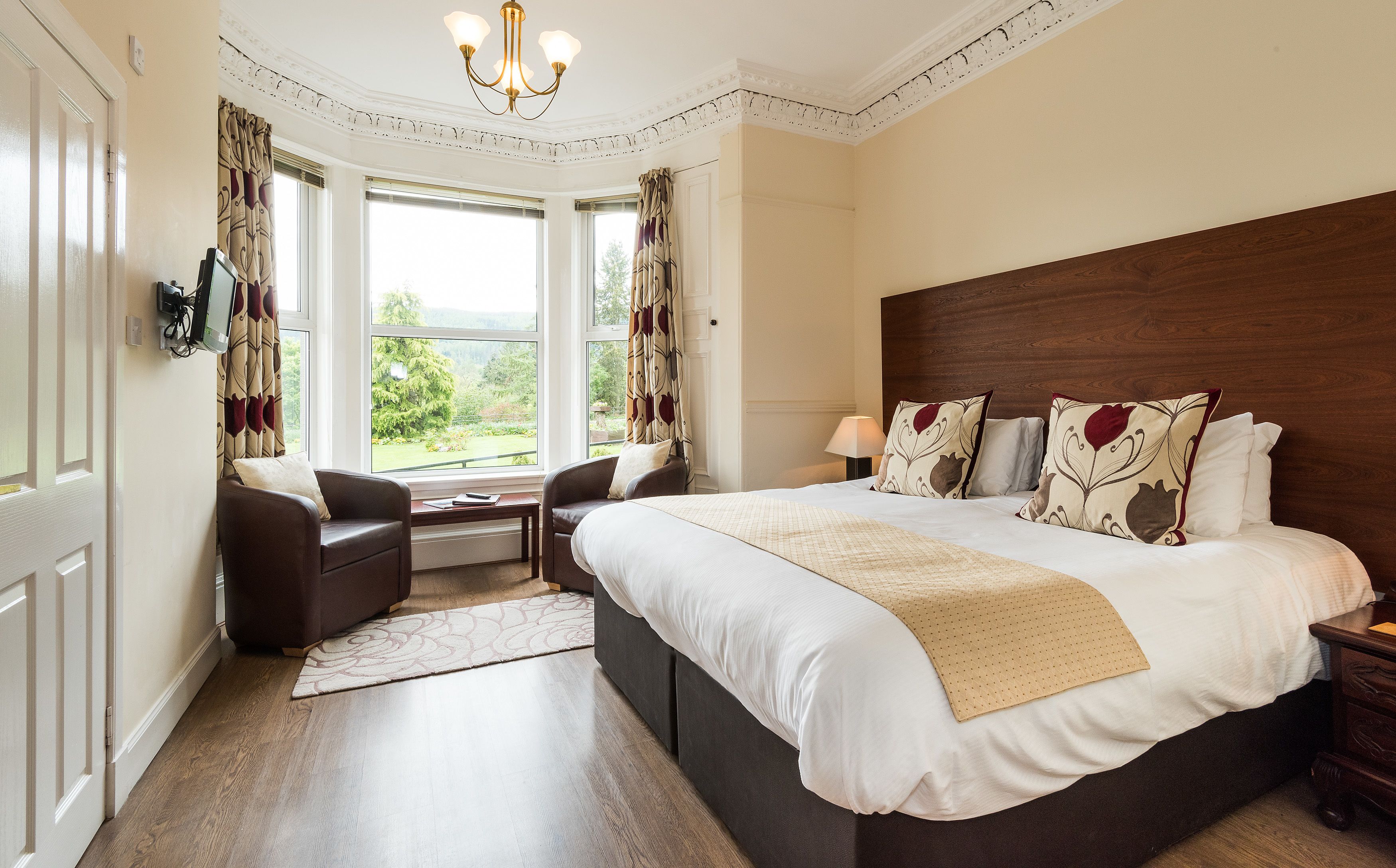 A hotel room with a large bed, two armchairs, a small table, and a view of the outdoors through large windows.