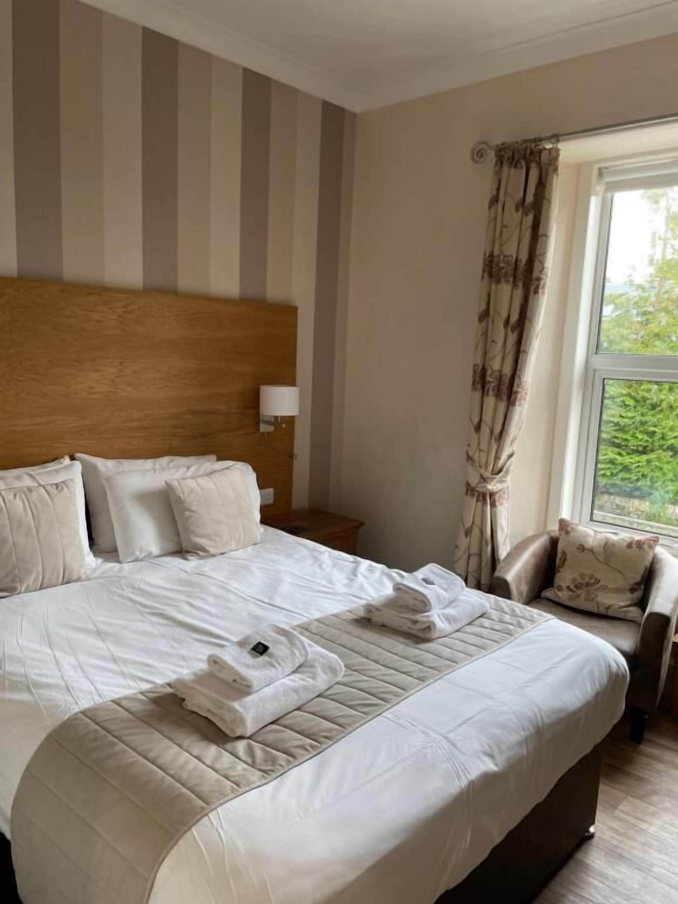 A hotel room with a large bed, a wooden headboard, a nightstand, a lamp, a chair, and a window with curtains.