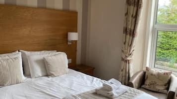 A hotel room with a large bed, a wooden headboard, a nightstand, a lamp, a chair, and a window with curtains.