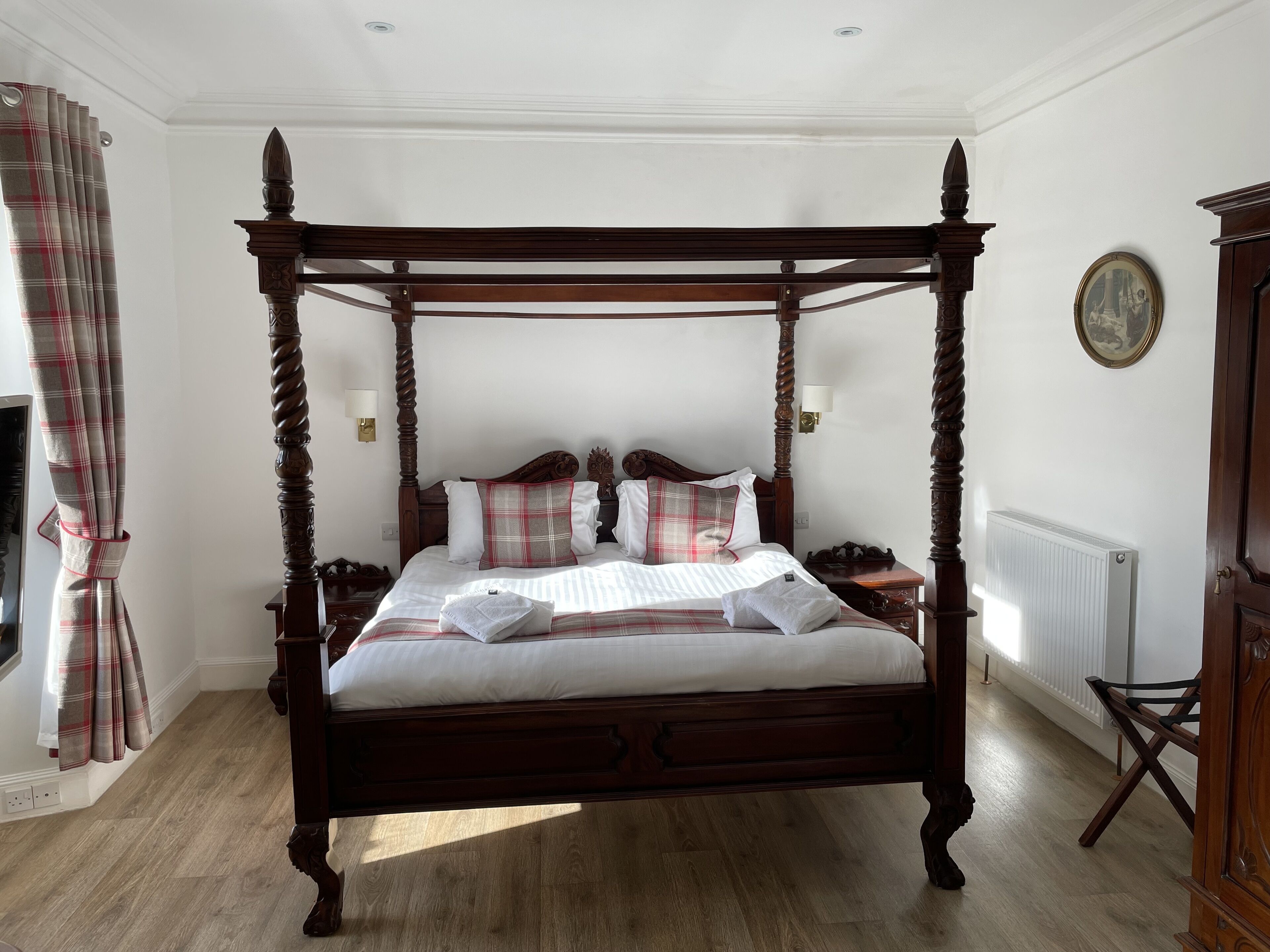 A four-poster bed with a wooden frame, white bedding, and plaid-patterned pillows. There is a wall-mounted light fixture and a radiator on the wall.